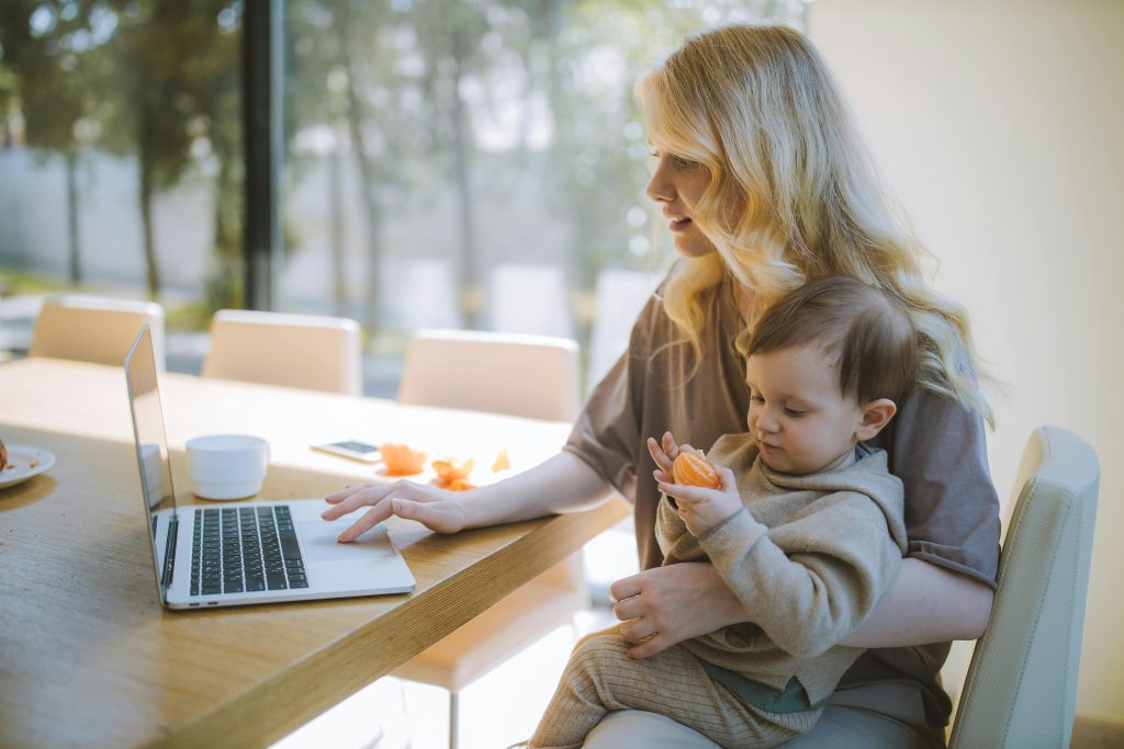 A woman working from home, holding her baby while typing on a laptop. The setting is relaxed, with the woman multitasking between work and childcare, demonstrating a blend of professional and personal responsibilities