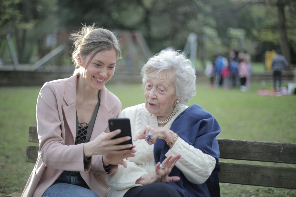 A cheerful senior woman and her adult daughter sit together, both engaged with a smartphone. They share a moment of joy and connection, smiling at each other while exploring the device.