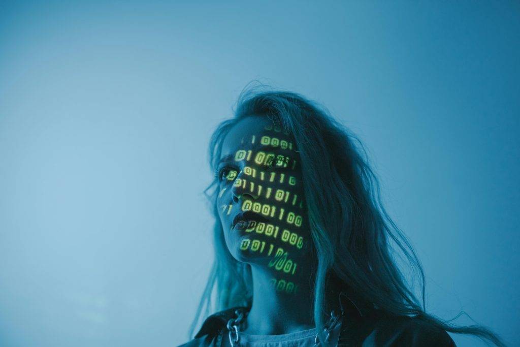 The image shows a person with long hair, bathed in cool blue lighting, giving the scene a futuristic, digital feel. Green binary code (ones and zeros) is projected onto their face, creating a blend of human and technology. The person appears thoughtful or introspective, gazing forward with a serious expression. The binary pattern on their face symbolizes the interaction between humans and the digital world, suggesting themes of technology, identity, or cybernetics.